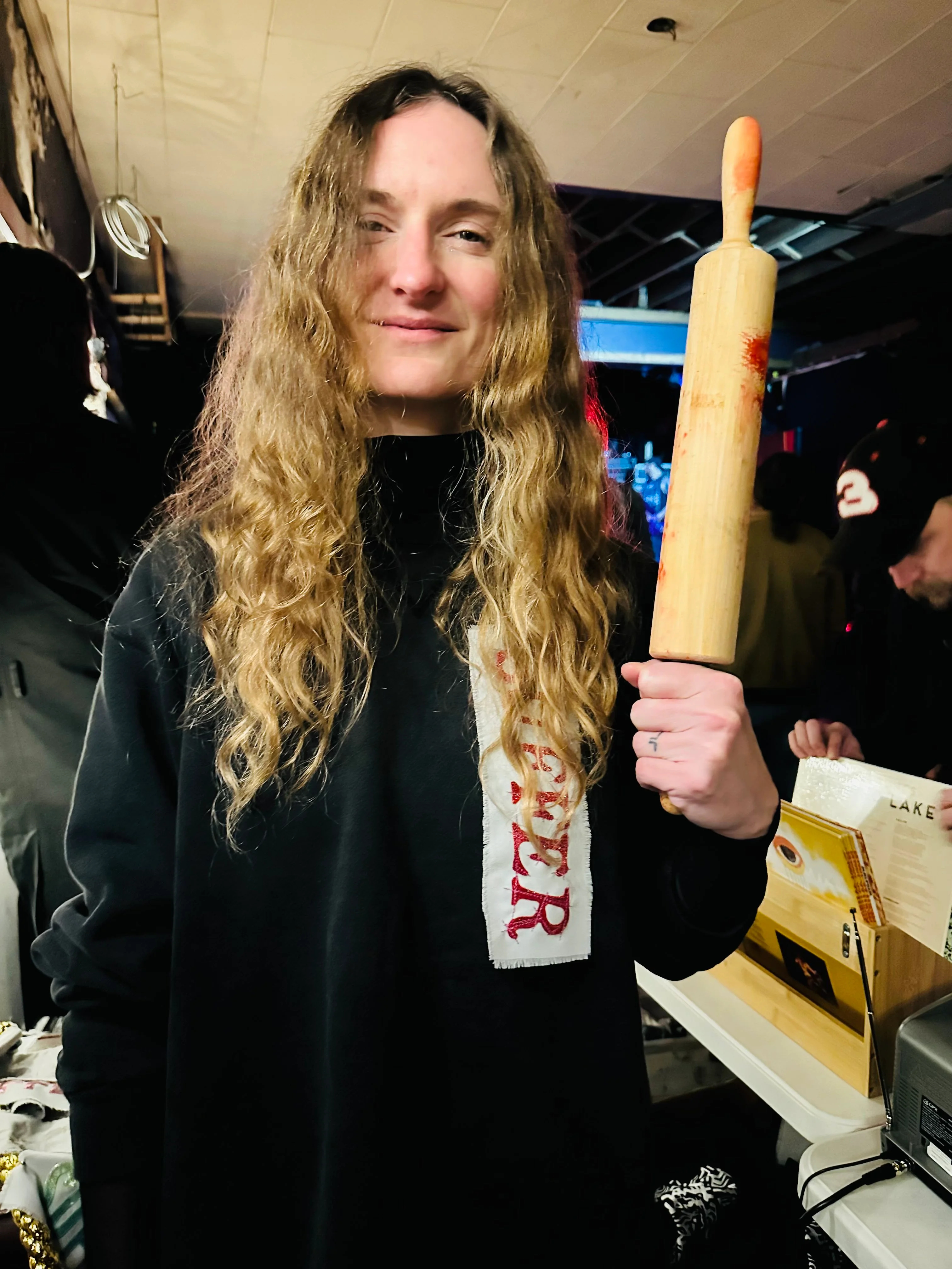 Candid photo of a woman who looks very tired holding a rolling pin, with a piece of fabric that says 'SUFFER' pinned to her sweatshirt.